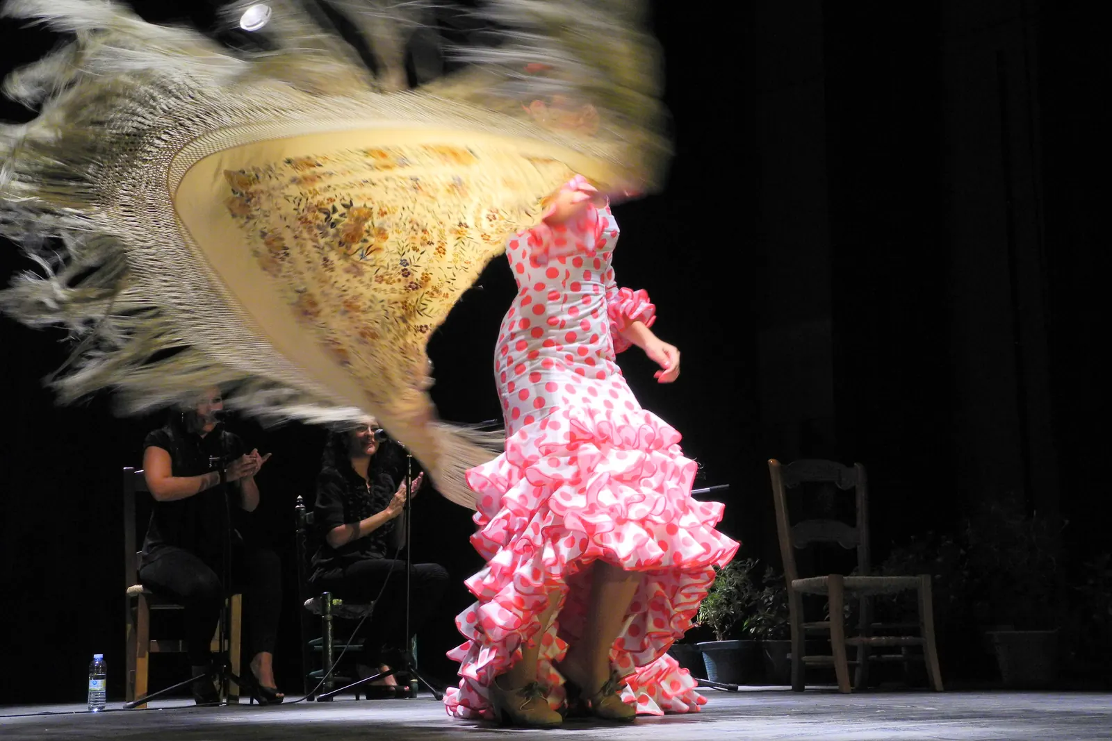 A flamenco dancer in a pink polka-dot dress spinning on a dark stage with a cream fringed shawl swirling around her