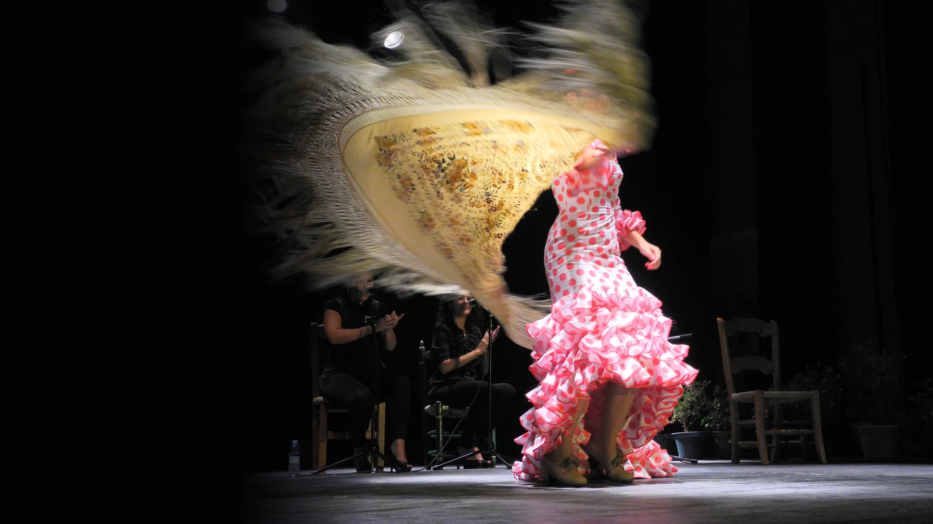 A flamenco dancer in a pink polka-dot dress spinning on a dark stage with a cream fringed shawl swirling around her, two seated women clapping behind