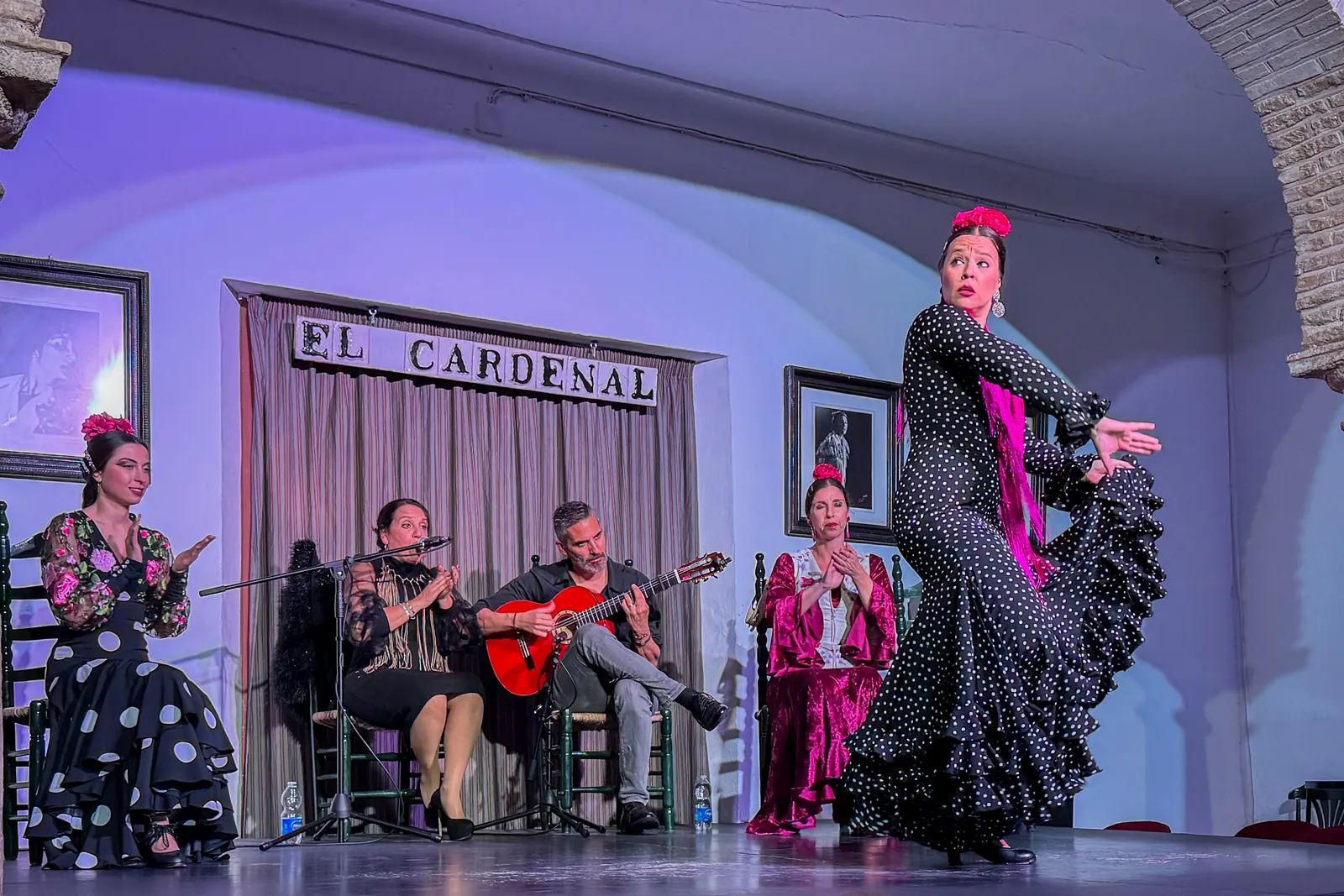 On a tablao stage a dancer in black with white spots turns while a singer, a guitarist and another dancer sit behind her